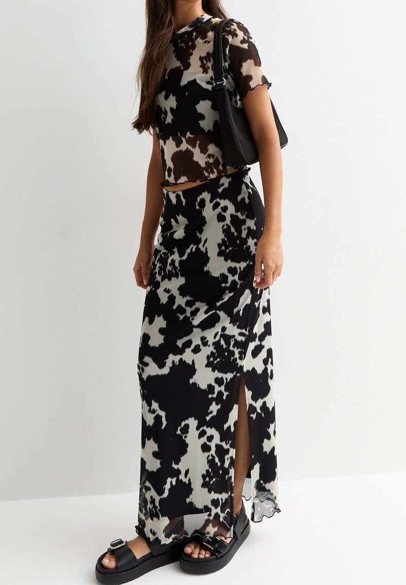 Femme portant un haut transparent imprimé vache noir et blanc et une jupe longue assortie avec une fente sur le côté, portant un sac à bandoulière noir et des sandales noires.