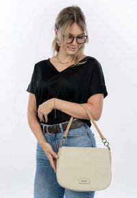 Borsa a tracolla in pelle di colore crema con una forma curvata, tracolla corta e accessori in argento, indossata con una maglietta nera e jeans blu.