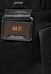 Detail van een zwarte tas van gestructureerde stof met een leren label waarop met reliëf "M.P." en het "Piquadro"-logo in metalen accenten staat.