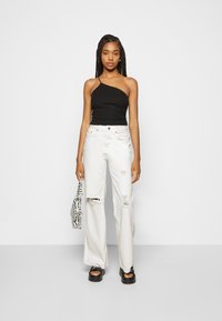 Top preto de um ombro só, jeans brancos largos e rasgados, e sandálias chunky pretas; segurando uma mala estampada de zebra. Tecido suave, design casual.