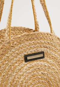 Sac rond en paille tissée avec des accents dorés, doté de poignées tressées et d'une étiquette noire. Des fibres naturelles texturées créent un motif rayé.