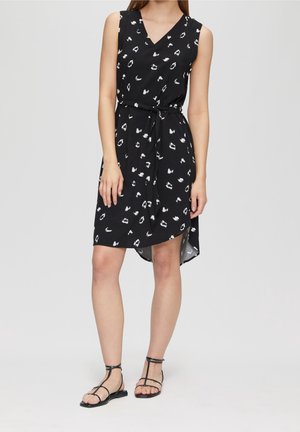 Kobieta w czarnej sukience bez rękawów z białymi abstrakcyjnymi plamami, dekoltem w serek, paskiem w talii i czarnych płaskich sandałach, stojąca na jednolitym tle.