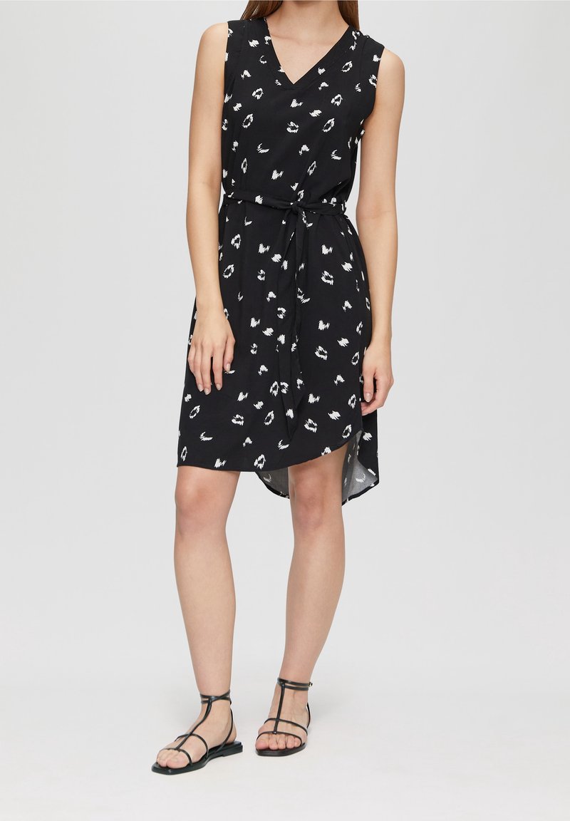 Femme portant une robe noire sans manches avec des taches abstraites blanches, col en V, ceinture à la taille, et sandales plates noires, debout devant un fond uni.