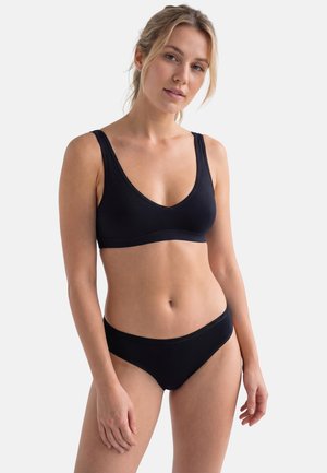 Donna che indossa un reggiseno nero e mutandine nere abbinate, in piedi contro uno sfondo chiaro e semplice, rivolta leggermente verso destra.