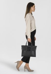 Zwarte leren tote bag met een textuur op het voorpaneel, dubbele handvatten en een slank rechthoekig ontwerp. Draagt een persoon in strak passende zwarte broeken en hakken.