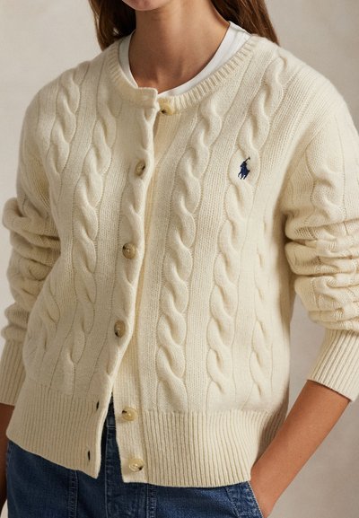 Femeie care poartă un cardigan crem tricotat cu model torsadat, cu nasturi, mâneci suflecate și logo brodat albastru închis pe piept, peste o cămașă albă.