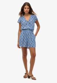 Romper de estampado floral azul con mangas cortas y escote en V. Tiene una cinturilla elástica y bolsillos laterales, combinado con sandalias de tacón marrones.