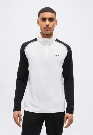 Mees, kes kannab valget ja musta pika varrukaga pusa poolläbi tõmblukuga, seisab tavalise heleda tausta ees.