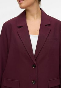 Bordeaux blazer med en klippet krave, enkelt knaplukning og en påsyet lomme på venstre side. Fremstillet af et glat, tekstureret stof.