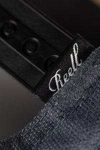 Dunkelgrauer Stoff mit strukturierter Oberfläche, versehen mit einem schwarzen Etikett mit weißer Schrift, auf dem "Reell" steht, und sichtbaren Hardware-Elementen im Hintergrund.