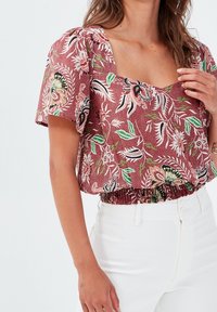 Rote florale Cropped-Bluse mit kurzen Puffärmeln, quadratischem Ausschnitt und gerafftem Saum. Mit grünen Blättern und schwarzen Akzenten. Kombiniert mit weißen Hosen.