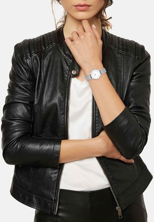 Veste de moto en cuir noir avec des détails côtelés, portée sur une chemise blanche ; montre à bracelet en maille argentée avec un cadran blanc au poignet.