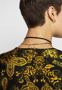 Czarna bluzka z żywym żółtym wzorem paisley. Zawiera zamek błyskawiczny z tyłu oraz warstwowe naszyjniki — jeden czarny wstążkowy i jeden złoty łańcuszek.