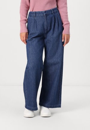 Person iført højtaljede, vidde blå denimjeans med plisseringer, kombineret med en lyserød langærmet sweater og hvide sneakers.