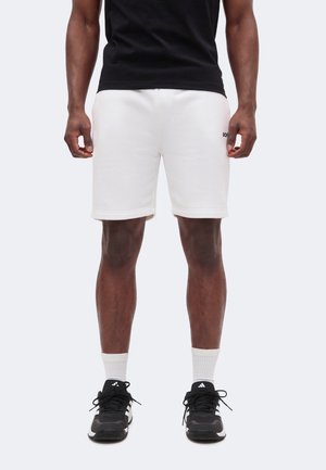 Uomo che indossa pantaloncini bianchi, t-shirt nera, calzini bianchi e sneakers nere Adidas, in piedi con le braccia rilassate lungo i fianchi.