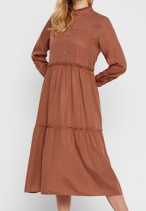 Nainen, joka on pukeutunut pitkähihainen, napillinen, kerroksittainen midi-mekko ruosteenruskeasta kankaasta, seisoo kädet selän takana.