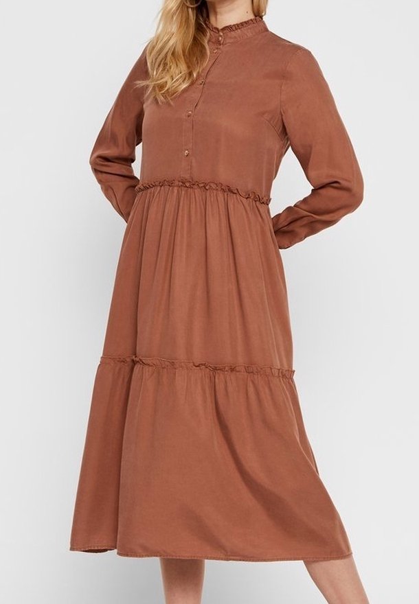Femme portant une robe midi à manches longues, boutonnée, à étages, en tissu brun rouille, debout les mains derrière le dos.
