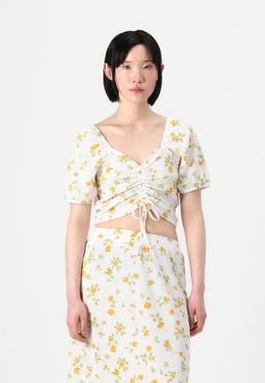 Hvit crop top med gult og grønt blomstermønster, med korte pufferm og rynketing foran, kombinert med et matchende skjørt.