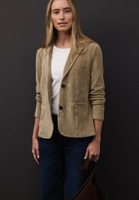 Femme portant un blazer en velours côtelé beige, une chemise blanche et un jean foncé, tenant un sac en cuir marron sur un fond sombre.