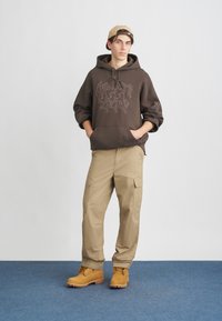 Felpa marrone con design grafico, pantaloni cargo beige con tasche laterali e stivali gialli con lacci e suola lavorata.