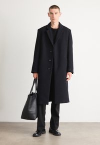 Abrigo de lana negro con un diseño de un solo botón, cierre de cinco botones y solapas amplias; combinado con pantalones negros y un bolso de piel.