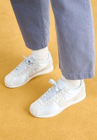 Sneaker bianche realizzate in suede e materiali sintetici, con una Swoosh grigia chiara, punta rotonda, lacci e suola in gomma testurizzata, abbinate a calzini beige e pantaloni blu.