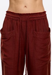Burgundy linen pants with an elastic waistband, drawstring, and two side pockets. Textured finish with gathered detailing at the waist.