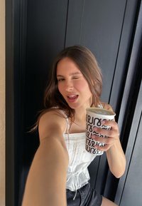 Top bianco testurizzato con lacci regolabili, pantaloni corti scuri, mentre tiene in mano una tazza a righe bianche e nere con arte del latte, sfondo con porta nera.