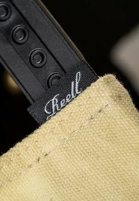 Yellow canvas fabric with a textured surface, featuring a black label with "Reell" in white script. Part of a black strap visible at the top.