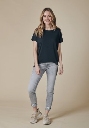 Smiling woman with long blonde hair stands wearing a black t-shirt, light gray rolled-up jeans, and beige sneakers on a plain background.