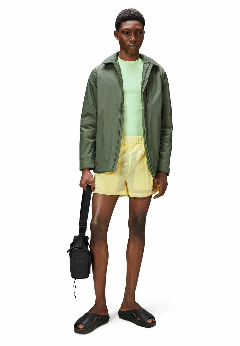 Veste verte résistante à l'eau, chemise vert clair, shorts jaunes, chaussures noires à enfiler et sac bandoulière noir.