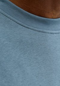 Mėlynas medvilnės audinys su sklandžia tekstūra, puoštas gofruota kaklo juostele. Artimas vaizdas rodo smulkių pynimo detalių.