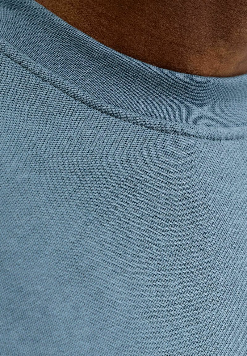 Mėlynas medvilnės audinys su sklandžia tekstūra, puoštas gofruota kaklo juostele. Artimas vaizdas rodo smulkių pynimo detalių.