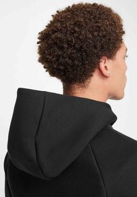 Sudadera negra con capucha ajustable, que presenta una textura suave y un tejido blando. Detalle de costura visible a lo largo de la costura del hombro.