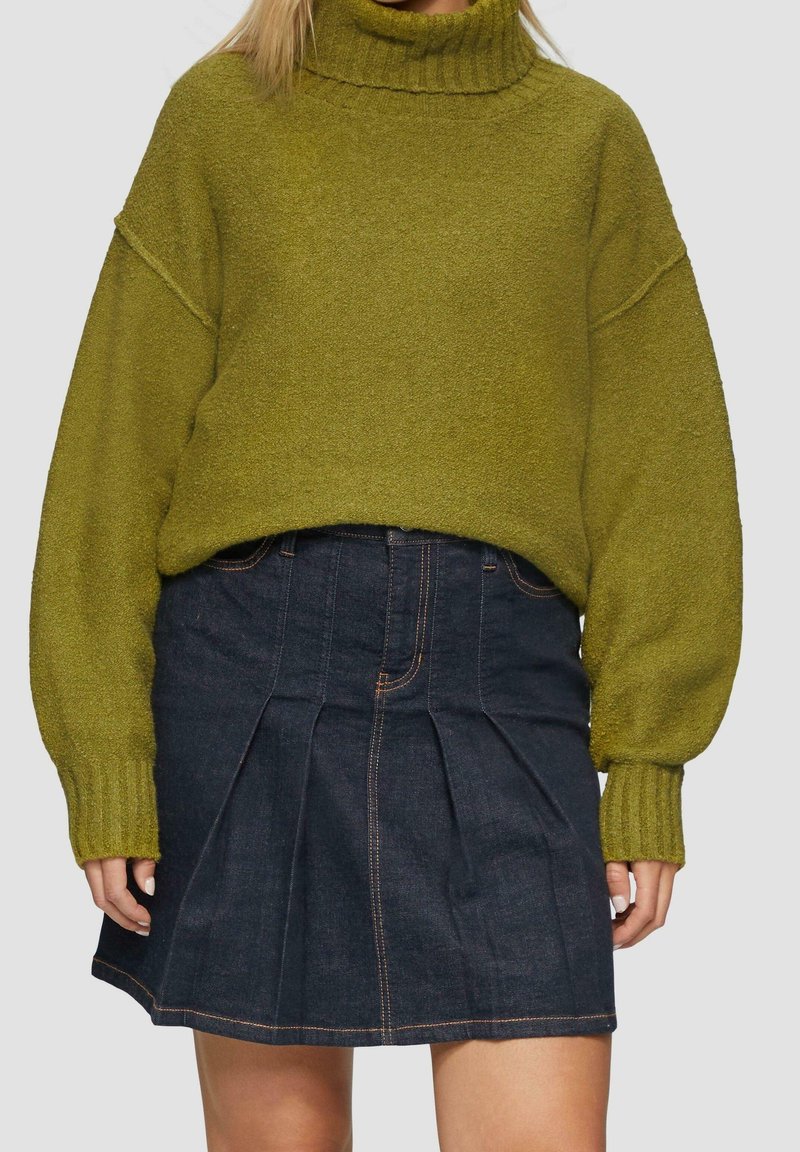 Femme portant un pull à col roulé en maille vert olive avec des manches ballon et une mini-jupe plissée en denim foncé.