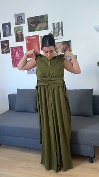 Γυναίκα που προσαρμόζει μακρύ ελιάς φόρεμα, στέκεται μπροστά από γκρι καναπέ με τοίχο γεμάτο διάφορα έργα τέχνης και φωτογραφίες πίσω της.