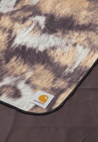 Dečka Carhartt s hnědým a krémovým vzorem zvířecího potisku, měkkou texturou a černým okrajem, obsahuje bílou logo nálepku.