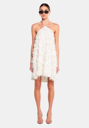 White halter dress featuring layered fringe texture, knee-length with a soft silhouette. Model wears black sunglasses and heeled sandals.