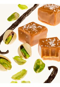 Carrés de caramel avec du gros sel, parsemés de pistaches et de gousses de vanille, sur fond clair. Les textures varient entre lisse et croquant.