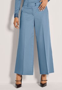 Vrouw in lichtblauwe hoge taille, wijde broek met voorplooien en zwarte puntige hakken, staande tegen een effen lichte muur.