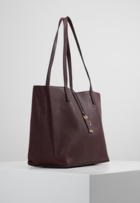 Borsa a tracolla in pelle bordeaux con due manici lunghi, una texture liscia e dettagli in metallo dorato. Presenta un design minimalista e una forma rettangolare.