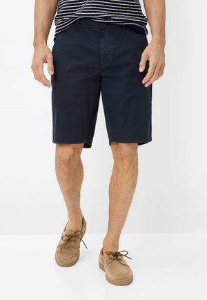 Man draagt marineblauwe shorts, gestreept shirt en beige casual veterschoenen, staand tegen een witte achtergrond.