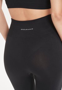 Zwarte high-waisted leggings van rekbare stof, met een gladde textuur en een cirkelvormige uitsnede aan de achterkant. Merken "ENDURANCE" op de tailleband.