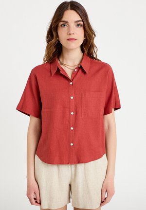 Kvinde med bølget brunt hår iført en kortærmet rustfarvet skjorte med knapper og lyse beige shorts, stående mod en ensfarvet hvid baggrund.