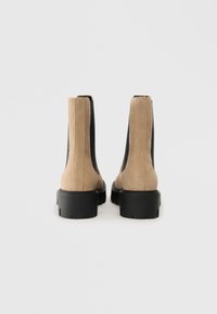 Beige suedestøvler til ankelen med svarte elastiske sider og kraftige svarte gummisåler. Har et rent design med teksturert overdel og glatt finish.