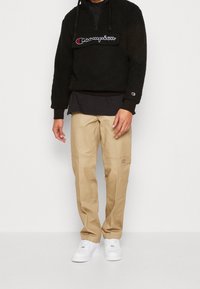 Schwarzer Fleece-Hoodie mit einem Frontreißverschluss und gesticktem Logo, kombiniert mit beigen, geraden Hosen und weißen Sneakers.