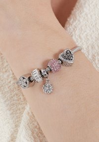 Silver charm bracelet featuring various beads: one pink enamel with rhinestones, heart charm, and flower-shaped pendant. Textured detail evident.