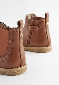 Botas de tobillo de cuero sintético marrón con paneles elásticos laterales, bordes festoneados, detalles perforados y una cremallera lateral. Suelas de goma tostada.