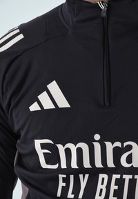 Camiseta deportiva negra con cremallera que presenta el logo blanco de Adidas y el texto "Emirates Fly Better", con una textura suave y lisa y un ajuste atlético.