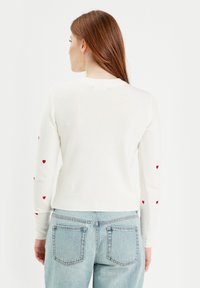 Suéter blanco de punto con adornos de corazones rojos en las mangas, con un cuello redondo y dobladillo acanalado, combinado con jeans de color azul claro.